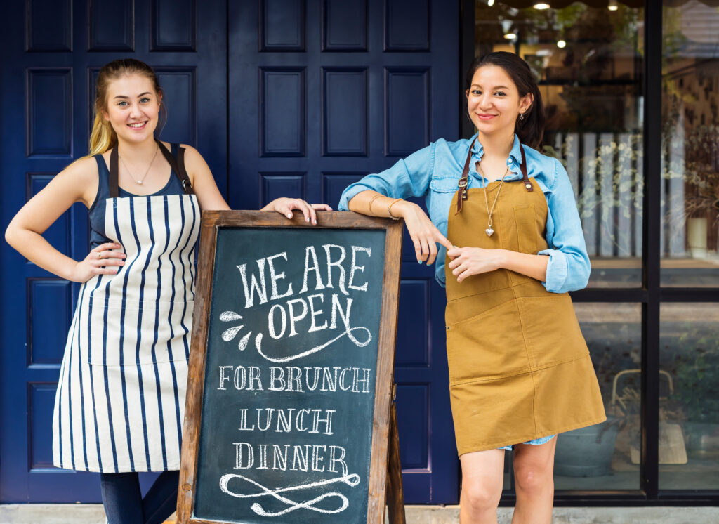 Cheerful,Business,Owners,Standing,With,Open,Blackboard