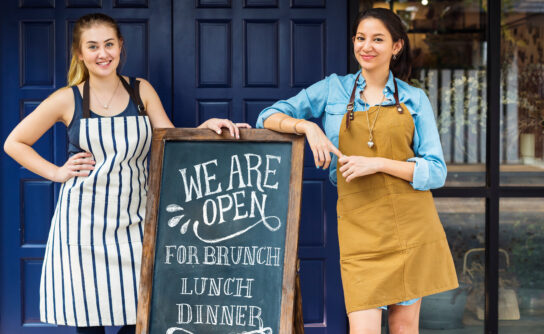 Cheerful,Business,Owners,Standing,With,Open,Blackboard